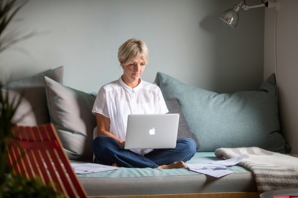 Kvinne med jeans, hvit bluse og kort lyst, hår sittende i sofa med laptop i fanget.