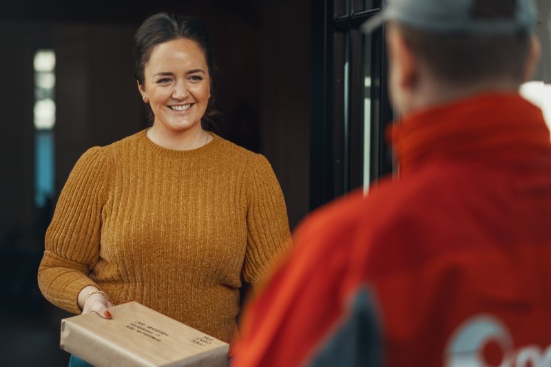Smilende kvinne med pakke i hånden som hun rekker mot et postbud.