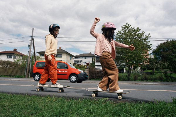 To barn på skateboard på et fortau. En rød bil fra Posten står parkert i bakgrunnen.