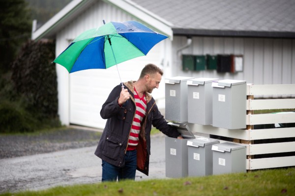 Mann med paraply henter post i postkasse plassert sammen med flere postkasser.
