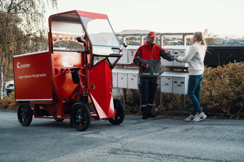 Postbud med elsykkel foran et postkassestativ. Leverer brev til en kvinne.