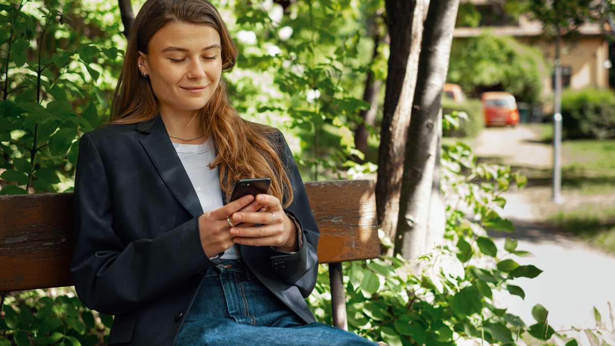 Ung kvinne som sitter på en benk foran et grønt tre og ser på mobilen sin