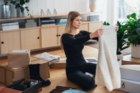 Kvinne som sitter på gulvet og holder en kjole foran seg. Bak henne ligger pakker og skoesker.