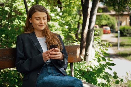 Ung kvinne som sitter på en benk foran et grønt tre og ser på mobilen sin