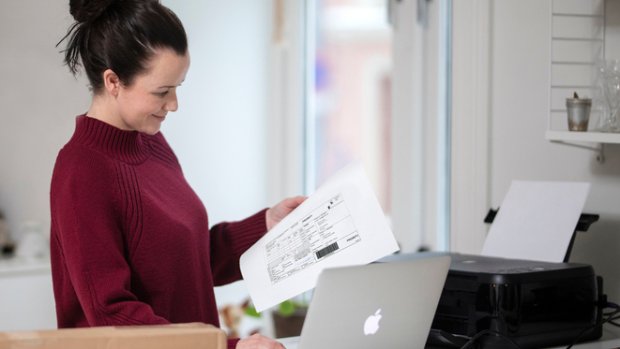 Dame med Mac og en skriver på benken og en utskrift av adresselapp med tollinformasjon i hånden.