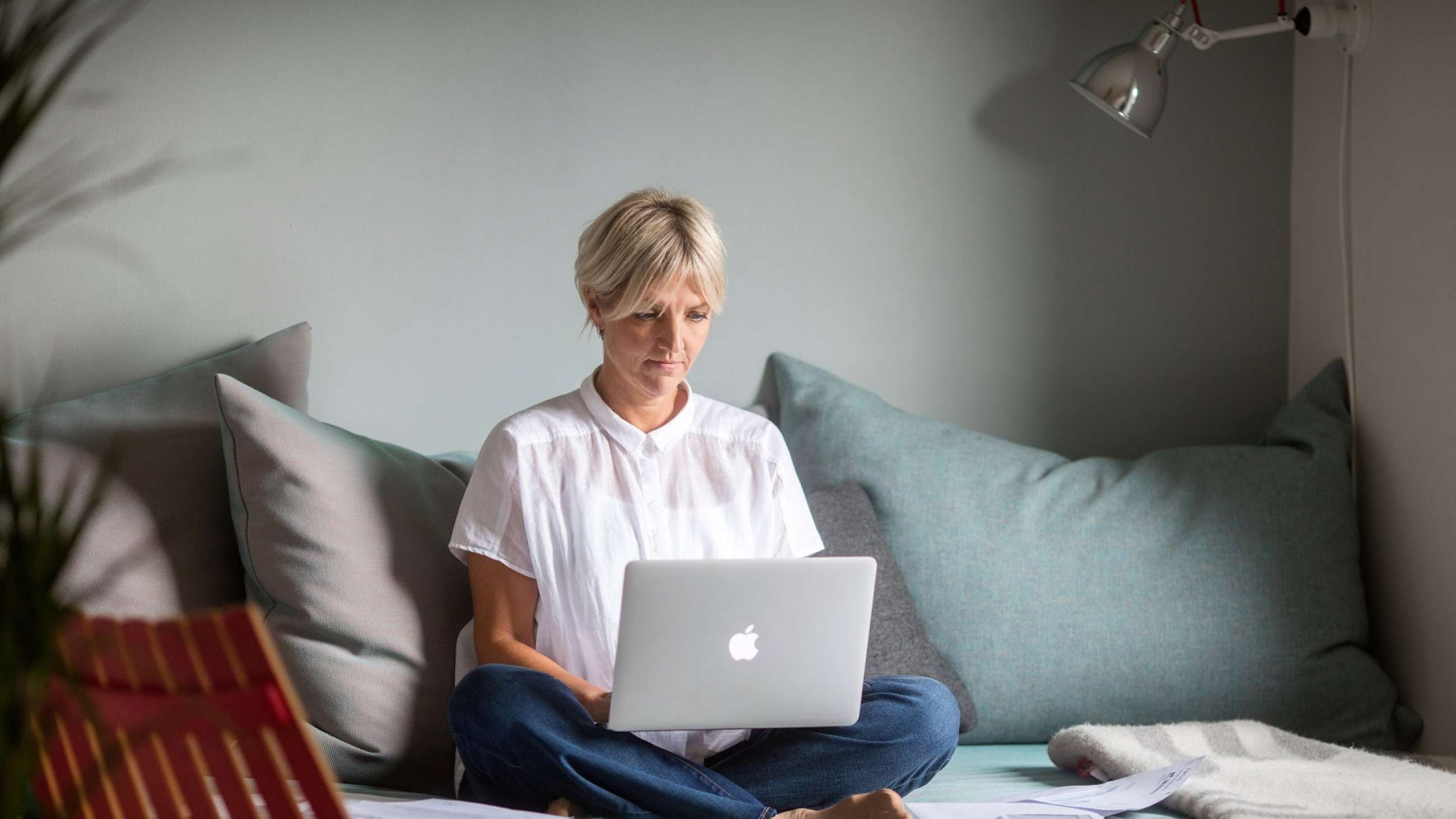 Kvinne med jeans, hvit bluse og kort lyst, hår sittende i sofa med laptop i fanget.
