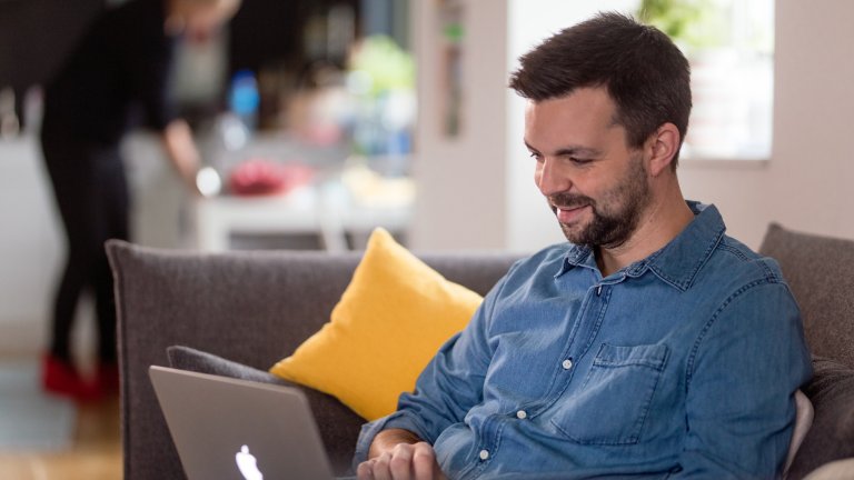 Mann med blå skjorte i sofa, sitter med en mac i fanget