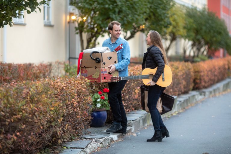 Ungt par på flyttefot. Han med pappesker og hun med en bag og gitar.