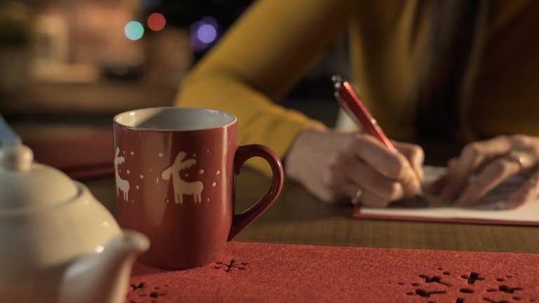 Kvinne sitter ved bord og skriver julekort. Kopp med julemotiv og tekanne i forkant av bildet.