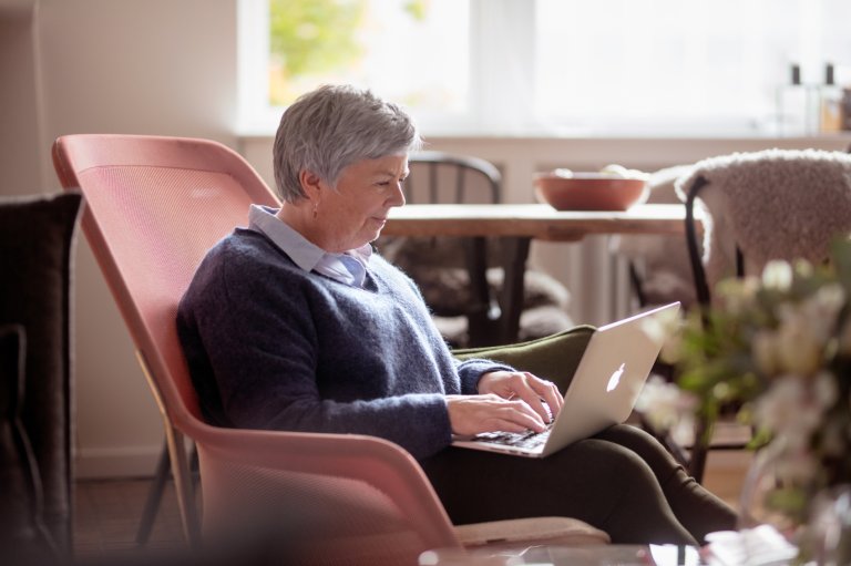 Eldre dame sitter i en rosa stol med en laptop på fanget