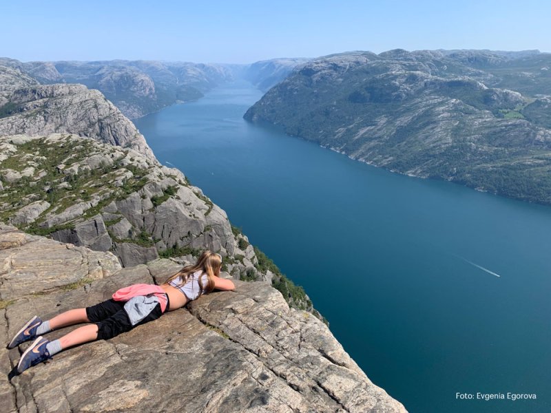 Jente ligger på kanten av et fjell og ser utover en fjord.