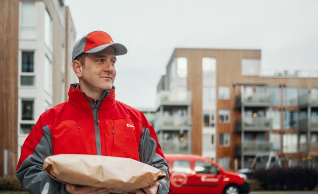 Postbud med pakke i hendene, ute i boligstrøk. Elektrisk postbil i bakgrunnen.